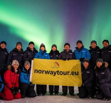 Travelers posing with a norwaytour.eu banner under the glowing Northern Lights during their Northern Lights tour from Svolvær.
