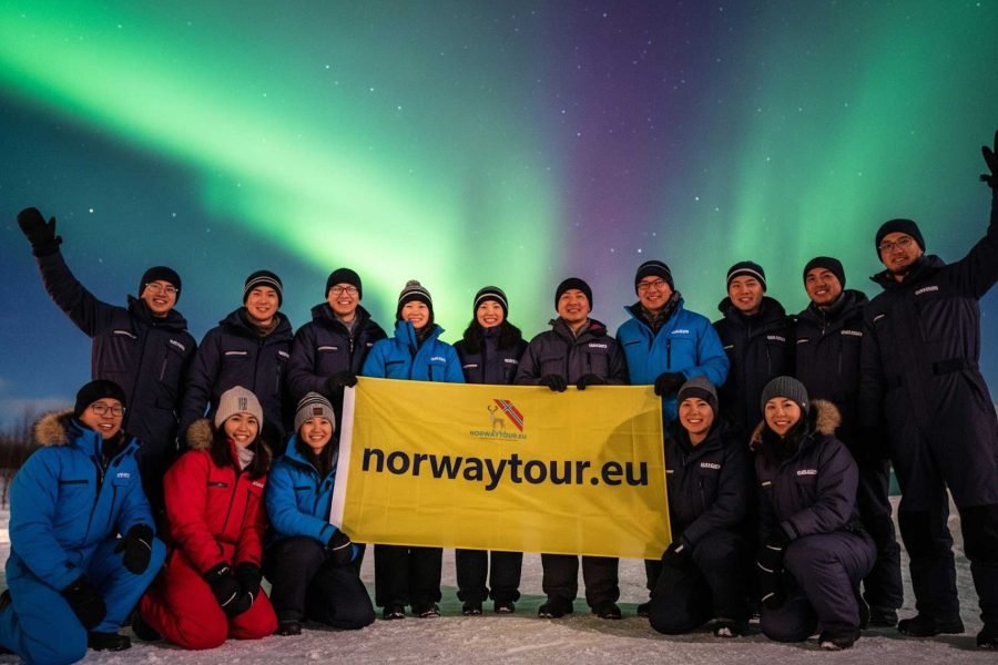 Travelers posing with a norwaytour.eu banner under the glowing Northern Lights during their Northern Lights tour from Svolvær.